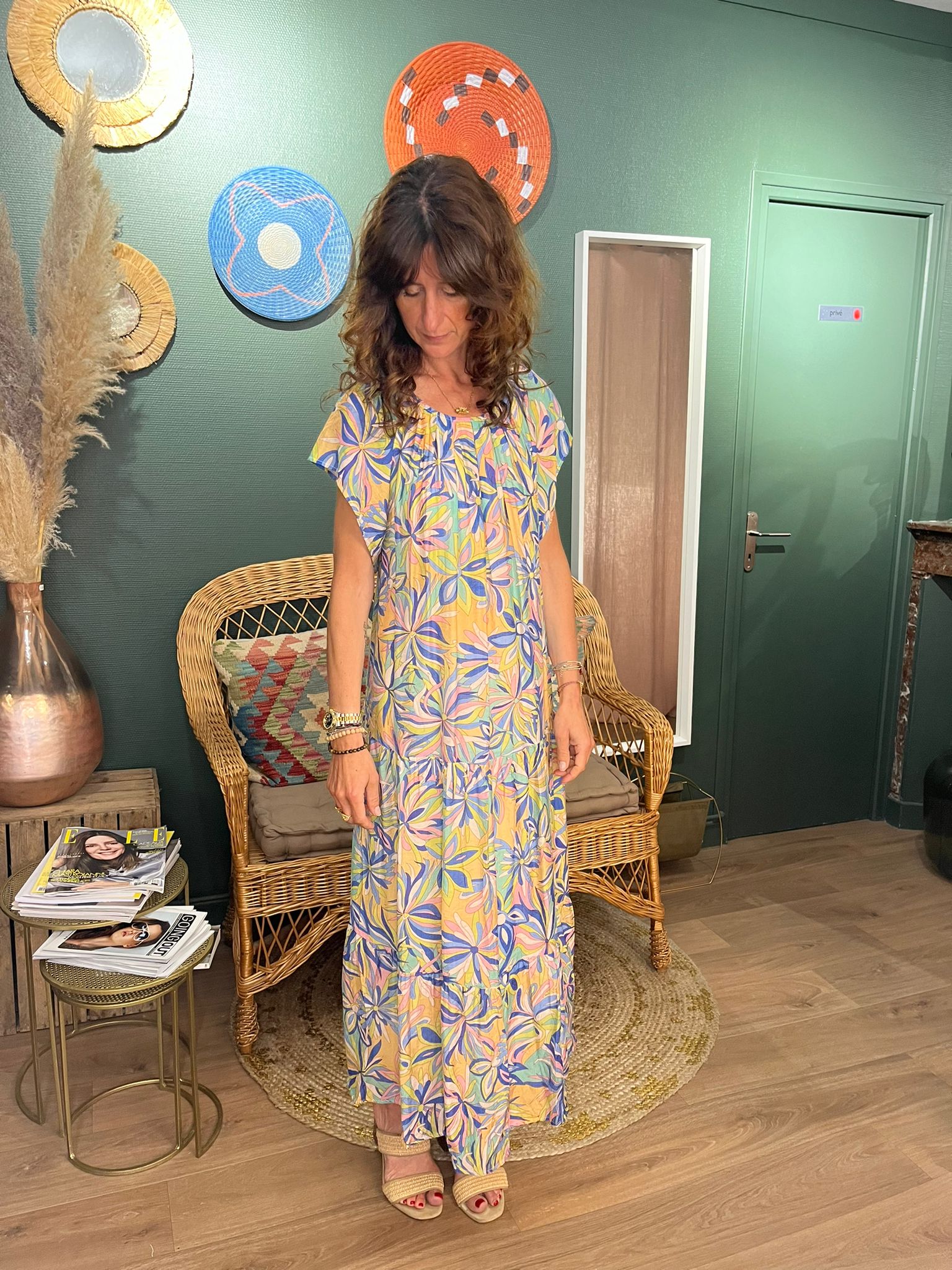 Alice portant la robe JULINE, une robe longue à motifs floraux multicolores, posant dans un intérieur boho-chic avec des décorations murales circulaires et des meubles en rotin.