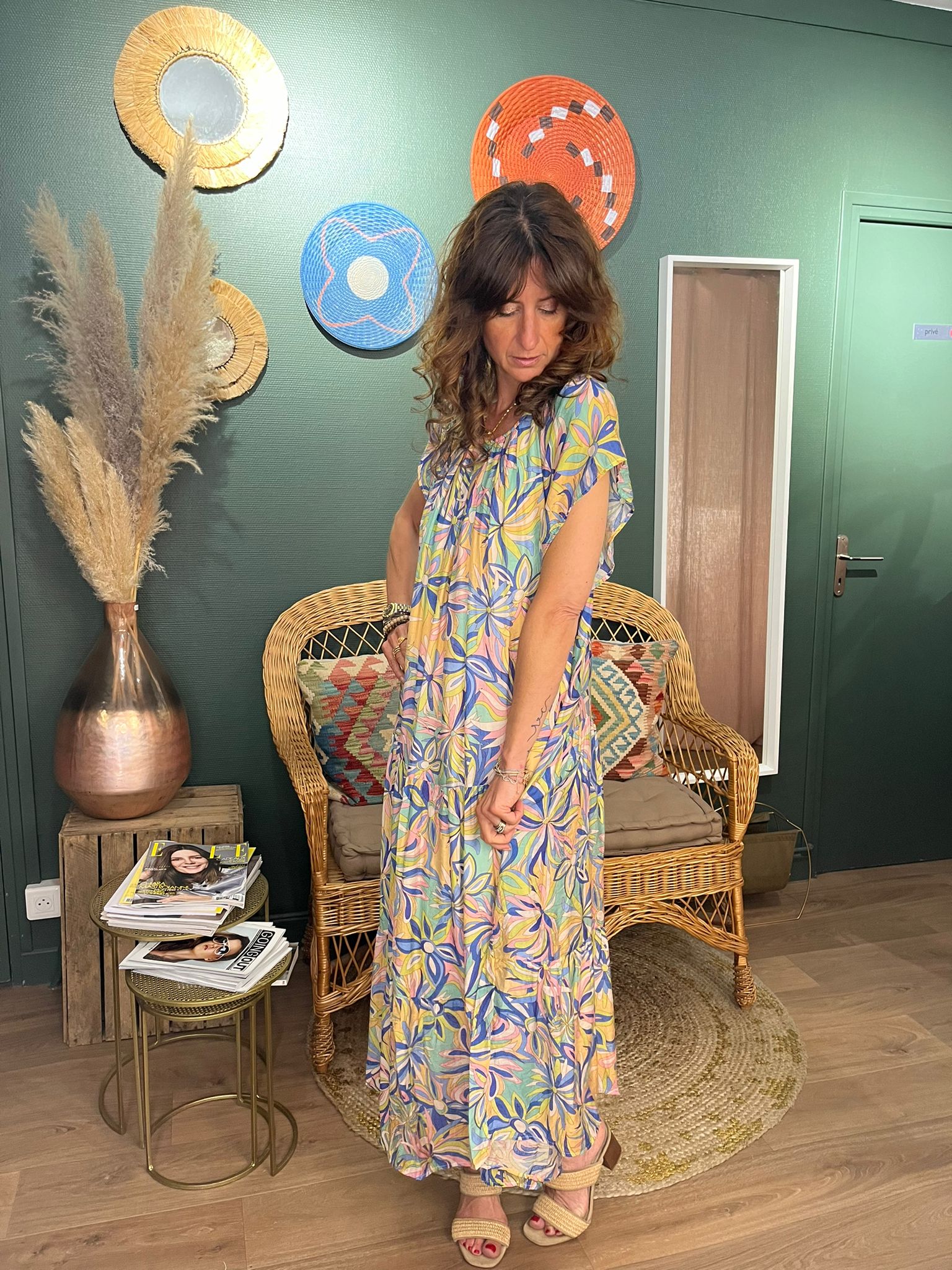 Alice portant la robe JULINE, robe longue à motifs floraux multicolores, regardant vers le bas dans un intérieur décoré avec des éléments boho-chic, assise sur une chaise en rotin.