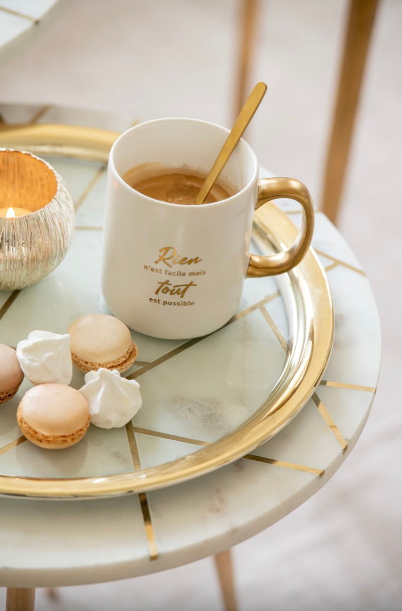 Mug en porcelaine blanche avec une anse dorée et l’inscription ‘Rien n’est facile mais tout est possible’ en doré, posé sur un plateau doré avec des macarons et des meringues