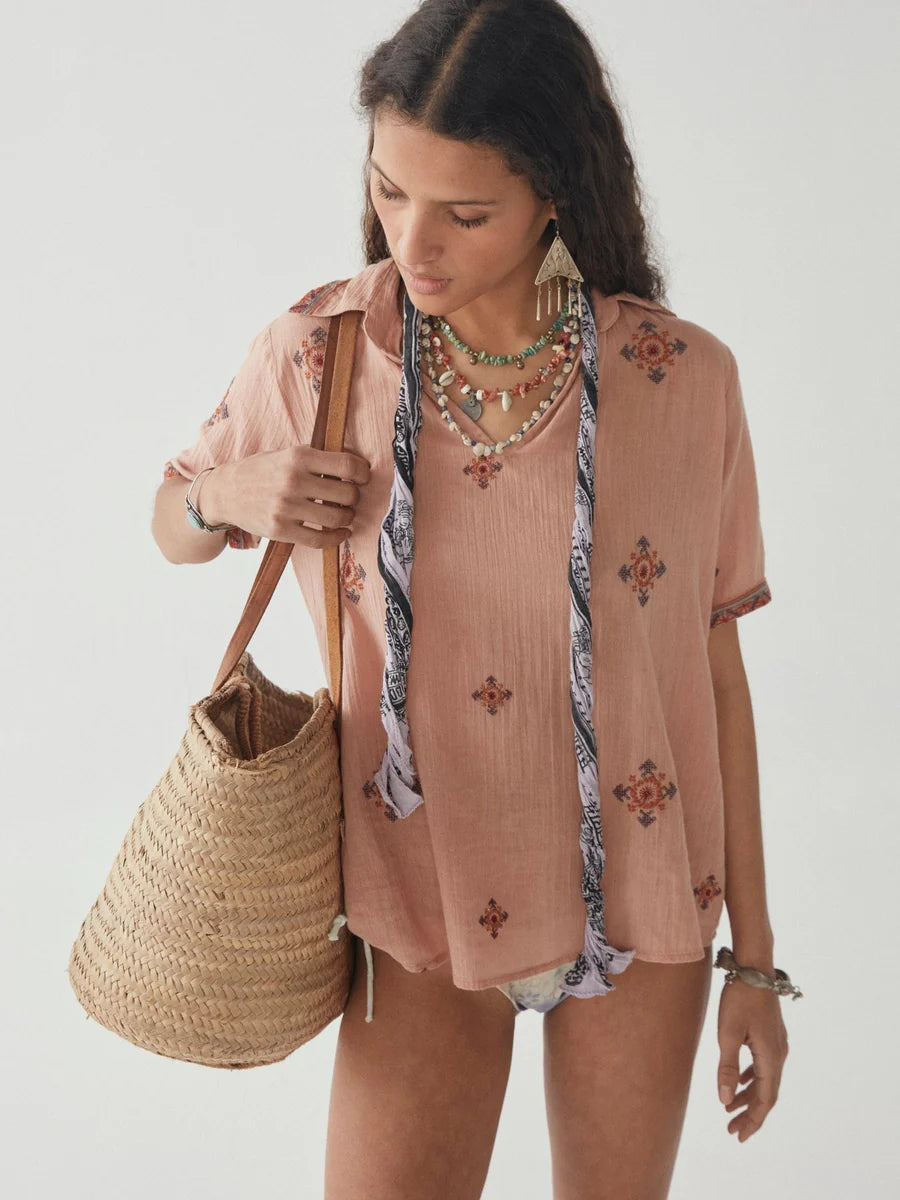 Femme portant la Blouse Allanis en couleur Compass Salmon avec un sac de plage en jute, montrant le style décontracté de la blouse à col en V et les broderies délicates. Vue de face.